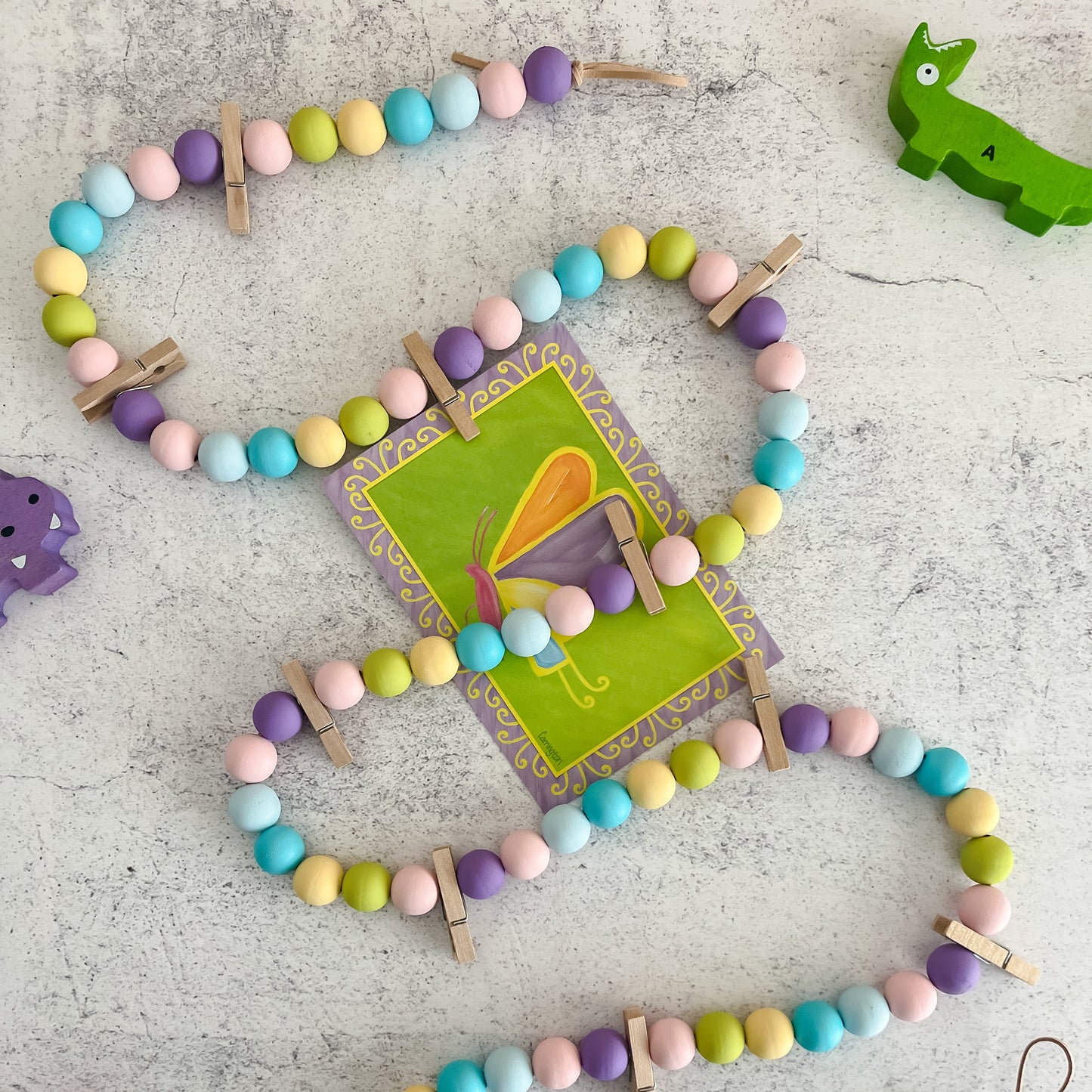 A colorful wooden bead garland with clothespins attached, designed for displaying artwork and photos, laid out on a grey surface.