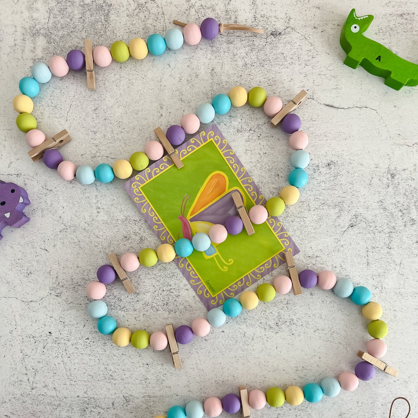 A colorful wooden bead garland with clothespins attached, designed for displaying artwork and photos, laid out on a grey surface.