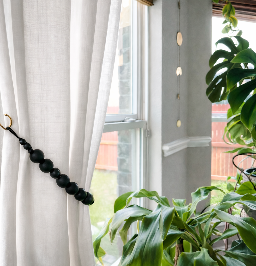 Black beaded curtain tieback on a white curtain with a window and plants in the background