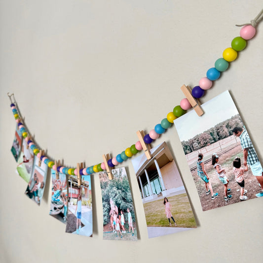 Rainbow photo garland with clothespins holding photos on a light gray wall.